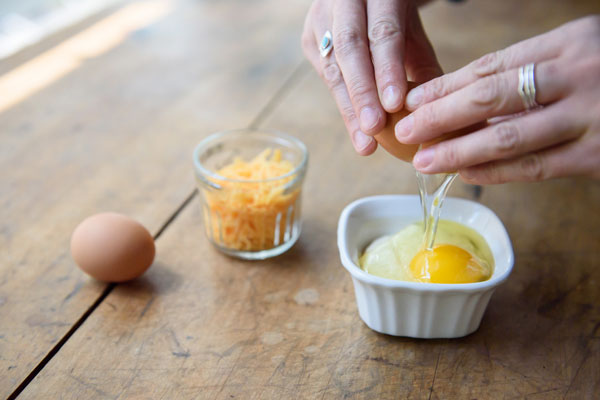 Woman's hands cracking egg into a ramekin on a wooden table with a glass of shredded cheese