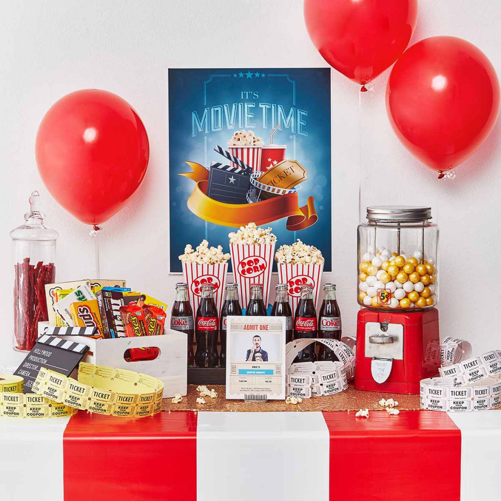 Table decorated with red and white striped tablecloth, gumball machine, bottles of Coca-Cola, candy, red balloons, tickets and popcorn