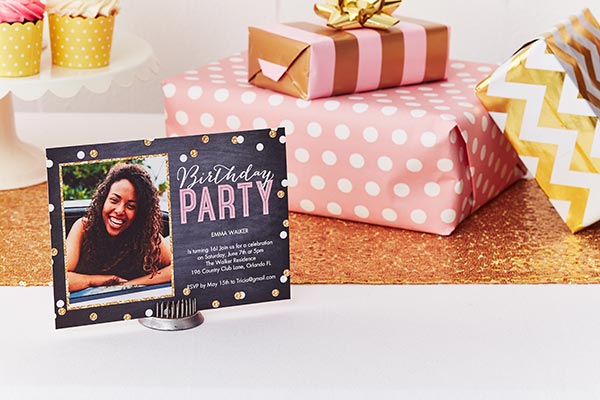 Sweet 16 birthday party photo invite with black background and gold and white dots, displayed on a table with pink and gold wrapped gifts